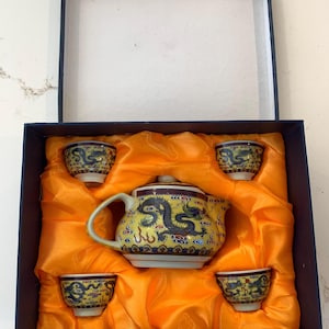 May include: A yellow ceramic tea set with a dragon design, including a teapot and four teacups, presented in a navy blue box with orange silk lining. The teapot and cups feature blue and red accents.