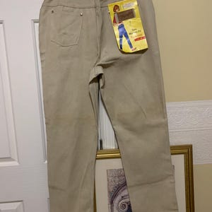 May include: Tan Wrangler jeans with a high waist and a straight leg. The jeans have a classic five-pocket design and a Wrangler tag attached. The tag features an illustration of a person wearing jeans. The jeans are hung on a hanger.