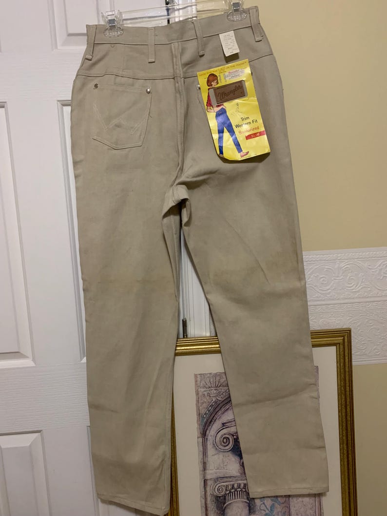 May include: Tan Wrangler jeans with a high waist and a straight leg. The jeans have a classic five-pocket design and a Wrangler tag attached. The tag features an illustration of a person wearing jeans. The jeans are hung on a hanger.