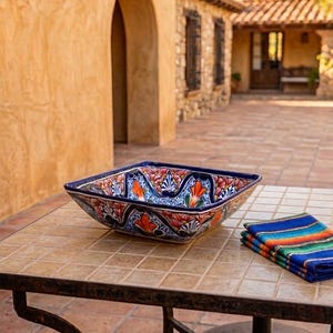 Puede incluir: Un cuenco cuadrado de cerámica con un vibrante diseño floral en naranja, azul y blanco se encuentra sobre una mesa embaldosada. El cuenco tiene un borde azul oscuro y está junto a un paño a rayas doblado. La mesa tiene una base de hierro forjado.