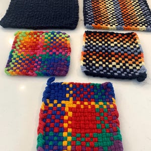 May include: A collection of five woven potholders in various colors and patterns. One is solid navy blue, while others feature stripes and checkerboard designs in orange, yellow, blue, green, and red. Each potholder is square-shaped and appears handmade.