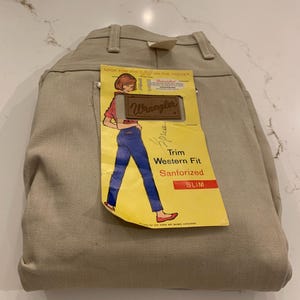 May include: Tan-colored Wrangler Slim Fit Western jeans. The jeans have a tag with a graphic of a woman in blue jeans and a red top. The tag reads "Trim Western Fit Sanforized SLIM".