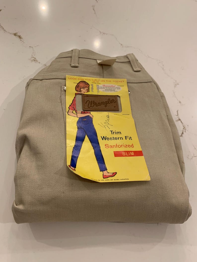May include: Tan-colored Wrangler Slim Fit Western jeans. The jeans have a tag with a graphic of a woman in blue jeans and a red top. The tag reads "Trim Western Fit Sanforized SLIM".