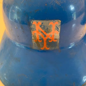 Op de afbeelding: Een vintage blauwe honkbalhelm met een oranje logo. De helm heeft een afgeronde bovenkant en een rand. Het logo is een gestileerd "NY"-ontwerp. De helm vertoont tekenen van slijtage.