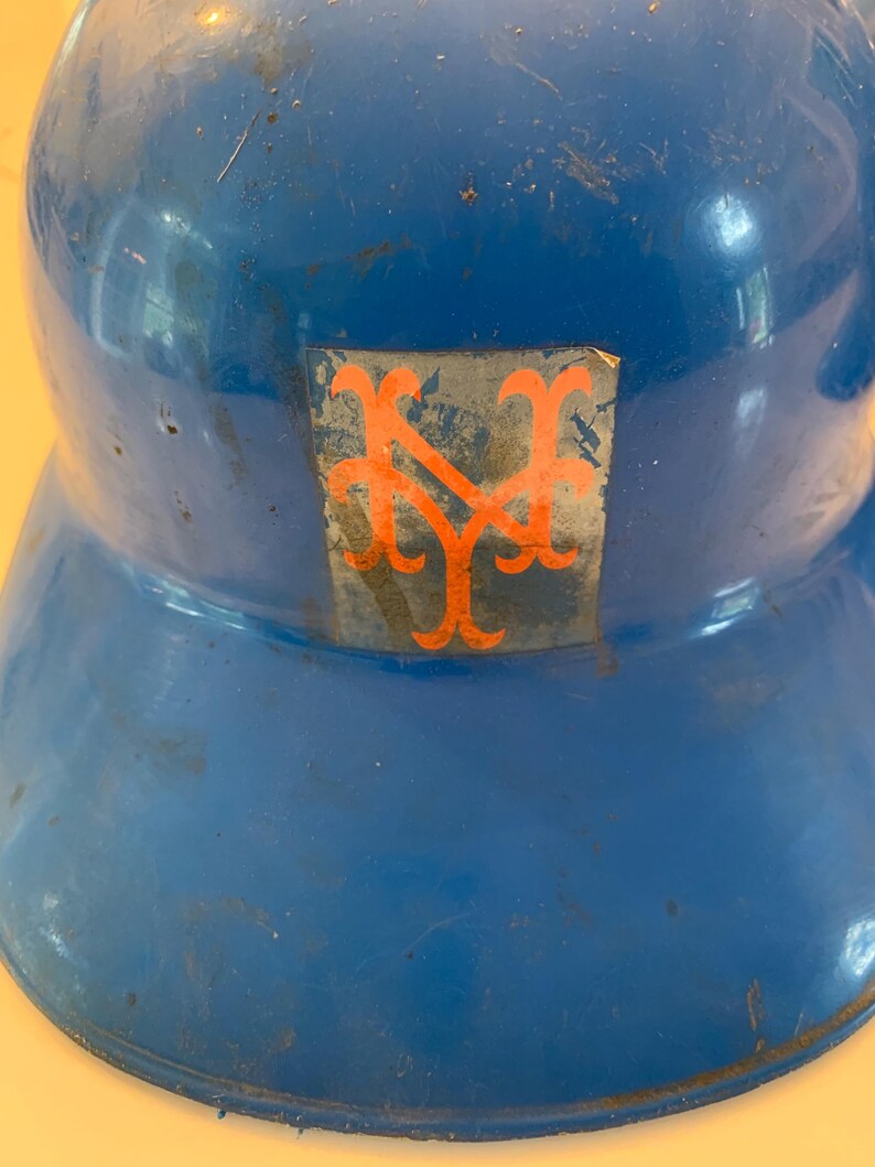 Op de afbeelding: Een vintage blauwe honkbalhelm met een oranje logo. De helm heeft een afgeronde bovenkant en een rand. Het logo is een gestileerd "NY"-ontwerp. De helm vertoont tekenen van slijtage.