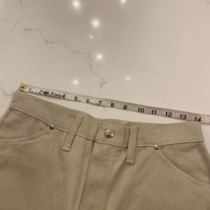 May include: Close-up of beige denim pants with a button closure and silver rivets. A measuring tape shows the waistline, with measurements in inches. The pants have classic five-pocket styling.