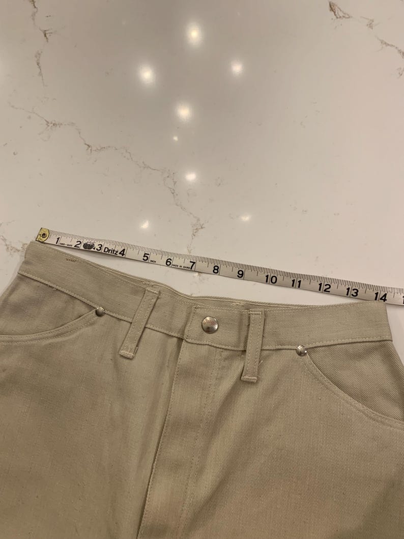 May include: Close-up of beige denim pants with a button closure and silver rivets. A measuring tape shows the waistline, with measurements in inches. The pants have classic five-pocket styling.