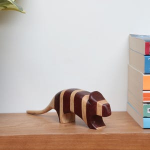 May include: A wooden animal figurine with brown and white stripes. The figurine is shaped like a small mammal with a long tail and a rounded head. It is sitting on a wooden shelf in front of a stack of books.