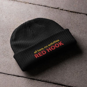 May include: A black knit beanie with the words "all bars, no activities" and "RED HOOK" embroidered in red and yellow thread. The beanie is folded at the brim and sits on a gray surface.