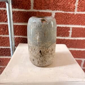 May include: A cylindrical ceramic vase featuring a two-tone glaze. The upper portion is a smooth, glossy light blue-grey, while the lower section has a textured, speckled beige and brown finish. The rim is rounded with a small, subtle notch.