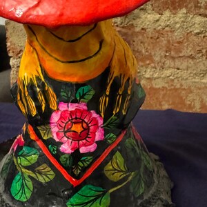 May include: A colorful paper mache figurine with a red hat, yellow scarf, and black and green floral details. The figurine is painted with bright colors and has a whimsical, folk art style.