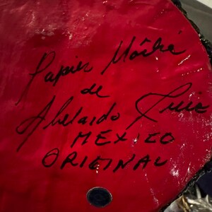 May include: Close-up of a red, round, ceramic object with black lettering. The text reads "Papel Mache de Ahelado Luis MEXICO ORIGINAL".