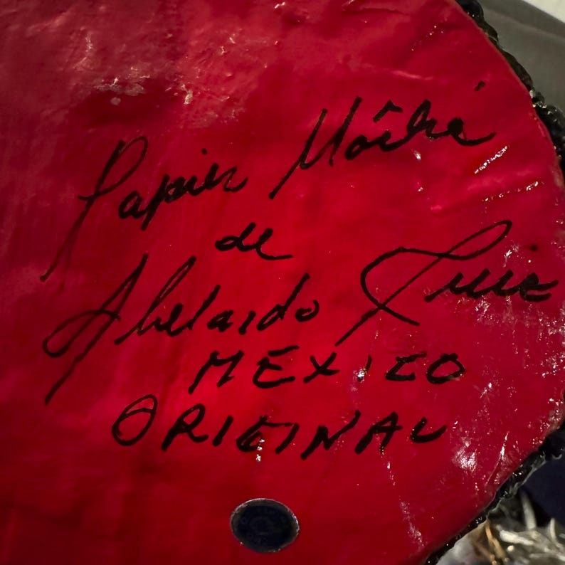 May include: Close-up of a red, round, ceramic object with black lettering. The text reads "Papel Mache de Ahelado Luis MEXICO ORIGINAL".