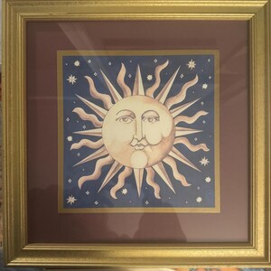May include: Framed artwork featuring a celestial sun design. The sun has a smiling face with radiating lines and is set against a dark blue background with stars. The artwork is framed in gold.
