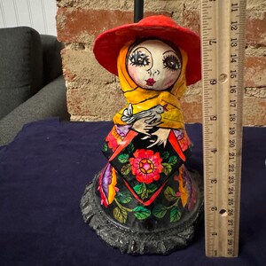 May include: A colorful paper mache figurine of a woman wearing a red hat, yellow scarf, and a black dress with floral designs. She is holding a white bird in her hands.