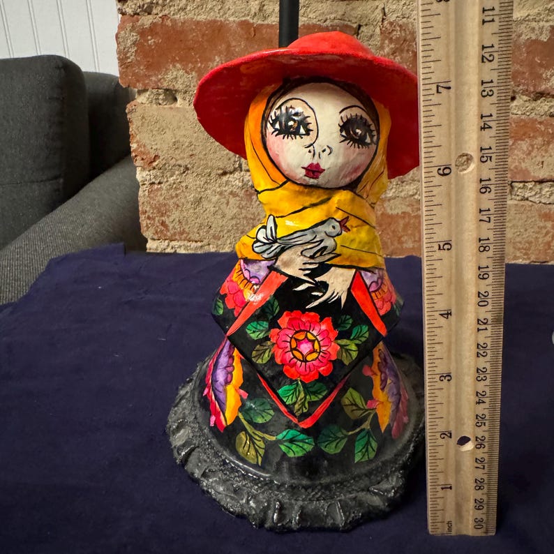 May include: A colorful paper mache figurine of a woman wearing a red hat, yellow scarf, and a black dress with floral designs. She is holding a white bird in her hands.