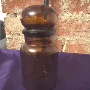 May include: A vintage amber glass apothecary jar with a round top and a black lid. The jar is cylindrical with a wide base and a narrower neck. The background is a brick wall and a purple surface.