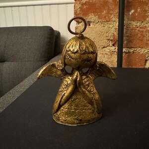 May include: A gold-colored angel figurine with closed eyes and folded hands in prayer. The angel has small wings and a halo-like ring at the top. The figurine is on a black surface.