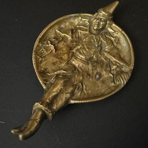 May include: A brass trinket tray shaped like a jester with a raised design. The jester is wearing a pointed hat and a jester's costume. The tray is oval shaped and has a smooth surface.