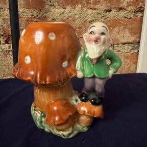 May include: A ceramic figurine of a gnome standing next to a mushroom planter. The gnome is wearing a green jacket, purple pants, and a white beard. The mushroom planter is brown with white polka dots.