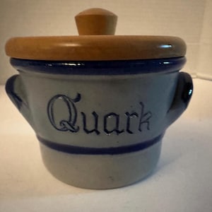 May include: A light gray ceramic crock with a dark blue rim and base, featuring the word Quark embossed in dark blue script on the front. The container has two side handles and a round, light brown wooden lid with a central knob handle.