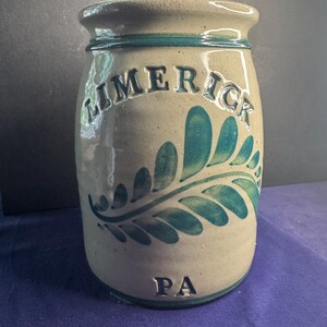 May include: A cylindrical stoneware crock with a beige base and a glossy finish. The vessel features a dark teal fern leaf design, the embossed text LIMERICK at the top, and the embossed text PA at the bottom, all in a matching dark teal color.