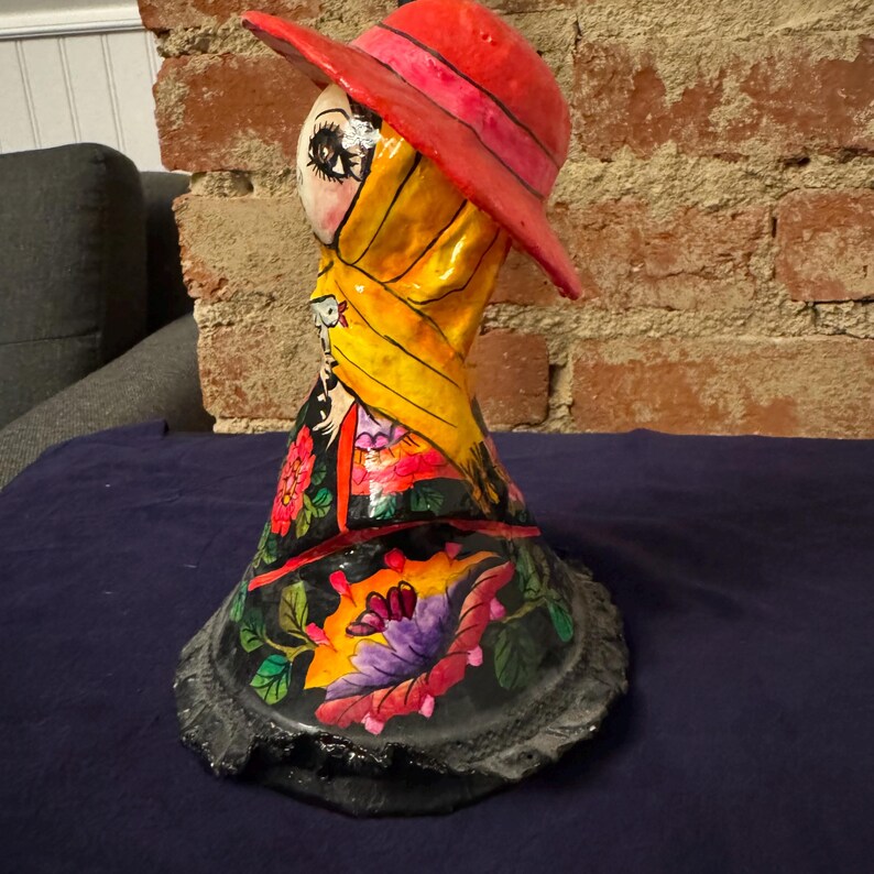 May include: A colorful paper mache figurine of a woman wearing a red hat and a yellow scarf. The figurine is decorated with flowers and other details.