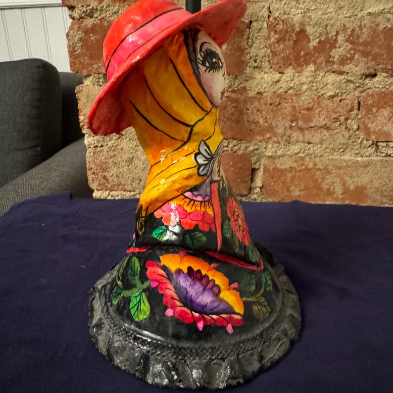 May include: A colorful paper mache figurine of a woman wearing a red hat, yellow scarf, and a floral dress. The figurine is painted with bright colors and has a detailed design.