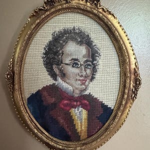 May include: An oval needlepoint portrait of a person with curly dark hair and glasses, wearing a dark coat, white ruffled collar, and a red bow tie. The artwork is encased in an ornate, metallic gold-toned oval frame with decorative detailing.