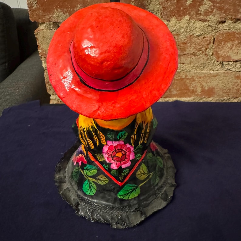 May include: A colorful paper mache figurine of a person wearing a red hat and a black and green patterned shawl with flowers. The figurine is sitting down and has a black base.