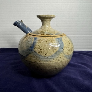 May include: A handmade ceramic teapot with a speckled beige and blue glaze. The teapot has a round body, a spout, and a lid. The spout and accents are a darker blue. The teapot is a unique piece of pottery.