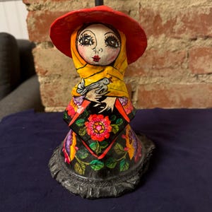 May include: A colorful paper mache figurine of a woman wearing a red hat, yellow scarf, and a black dress with floral designs. She is holding a white bird in her hands.
