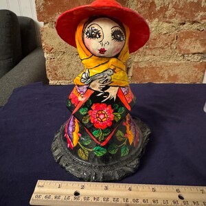May include: A colorful paper mache figurine of a woman wearing a red hat, yellow scarf, and a black dress with floral designs. She is holding a white bird in her hands.