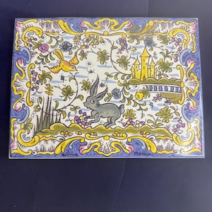 May include: Rectangular ceramic tile with a detailed, colorful design. The tile features a gray rabbit, yellow bird, and a yellow castle. The background includes floral and vine motifs in blue, yellow, and purple. The words "RUSTICA PORTUGAL" are at the bottom.