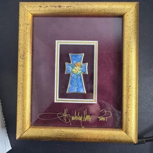 May include: Framed artwork featuring a blue cross with a gold border, depicting a figure in the center. The cross is set against a burgundy background within a gold-colored frame. The artwork is signed and dated 2001.
