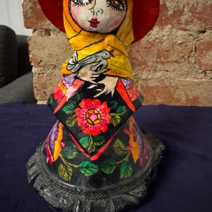May include: A colorful paper mache doll with a red hat, yellow scarf, and floral skirt. The doll is holding a white dove in its hands.