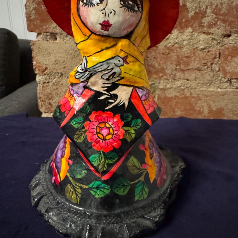 May include: A colorful paper mache doll with a red hat, yellow scarf, and floral skirt. The doll is holding a white dove in its hands.