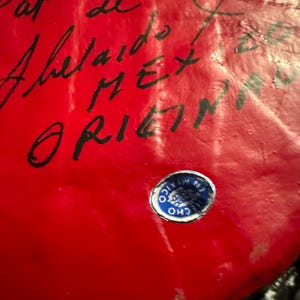 May include: A red, round object with black writing that says "Adelaide" and "MEX ORIGIN". A blue and silver sticker with a logo is on the object.