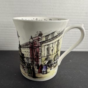 May include: White ceramic mug featuring a detailed illustration of a European street scene with buildings, people, and streetlights. The mug has a curved handle and a slightly flared rim. The artwork is in black, yellow, red, and purple.