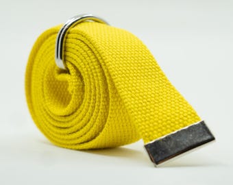 May include: A rolled-up yellow canvas belt with a silver-colored metal buckle. The belt has a rectangular metal end piece. The bright yellow color contrasts against the white background.