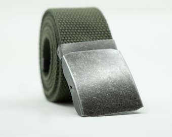 May include: An olive green canvas belt rolled into a circular shape, featuring a rectangular, brushed silver-tone metal buckle with a distressed finish. The belt strap has a visible woven texture and is positioned at an angle on a white surface.