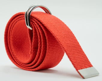 May include: A rolled-up red canvas belt with a silver-colored D-ring buckle. The belt's end has a silver-colored metal tip. The fabric has a textured weave. The belt is a fashion accessory.