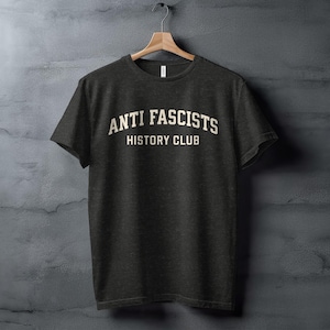 Puede incluir: Camiseta gris oscuro con el texto blanco « ANTI FASCISTS HISTORY CLUB ». La camiseta está colgada de una percha de madera contra una pared gris texturizada. Tiene cuello redondo y mangas cortas.