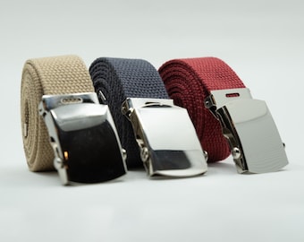May include: Three canvas belts in tan, navy blue, and red, each with a shiny silver-colored buckle. The belts are rolled up, showcasing their texture and color variations. The buckles are rectangular with a smooth, polished finish.