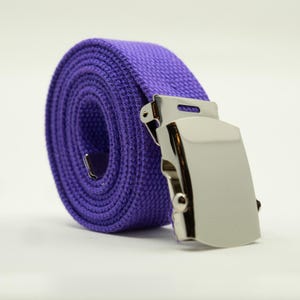 May include: A rolled-up purple canvas belt with a silver-colored metal buckle. The belt is made of a woven fabric and has a classic, casual design. The buckle is rectangular and has a shiny finish.