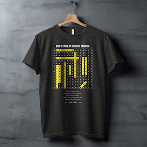 以下が含まれることがあります： 黄色と白のクロスワードパズルデザインの濃い灰色のTシャツ。「250 Years of Hidden Truths」と書かれたテキストと、下部にテキストが含まれています。木製のハンガーにTシャツが掛かっています。