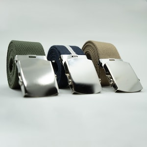 May include: Three canvas belts with silver-colored metal buckles. The belts are rolled up, showcasing their different colors: olive green, navy blue with a white stripe, and tan. The buckles are rectangular and shiny.