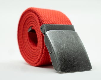 May include: A rolled-up red canvas belt with a silver-colored metal buckle. The belt is made of a woven fabric and has a simple, classic design. The buckle is rectangular and has a matte finish.