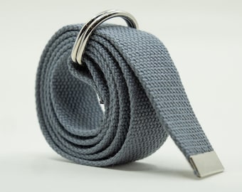 May include: A slate blue canvas belt rolled into a circular shape, featuring two polished silver-tone metal D-ring buckles at the top and a matching silver-tone metal tip at the end of the fabric strap, showcasing a textured woven material.