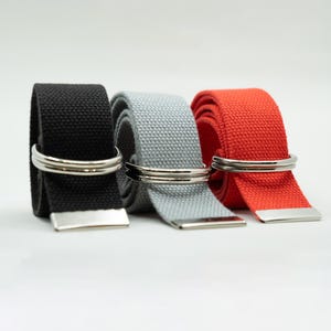 May include: Three woven fabric belts in black, light gray, and bright red. Each belt features a double silver-toned metal D-ring buckle and a matching silver-toned metal tip at the end of the strap, showcasing a textured, durable weave construction.